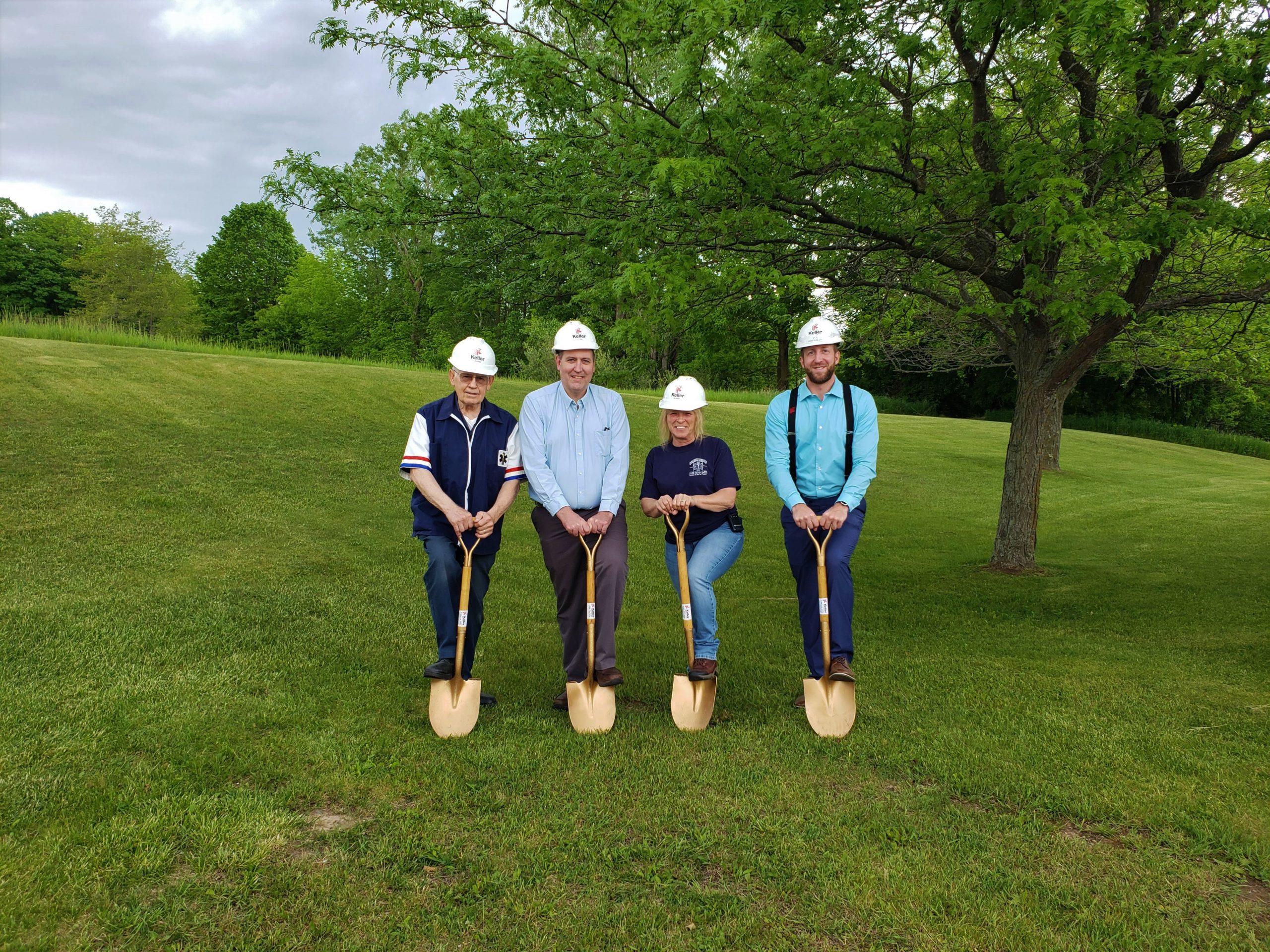 Team of four holding gold shovels and wearing hard hats at groundbreaking in grassy area