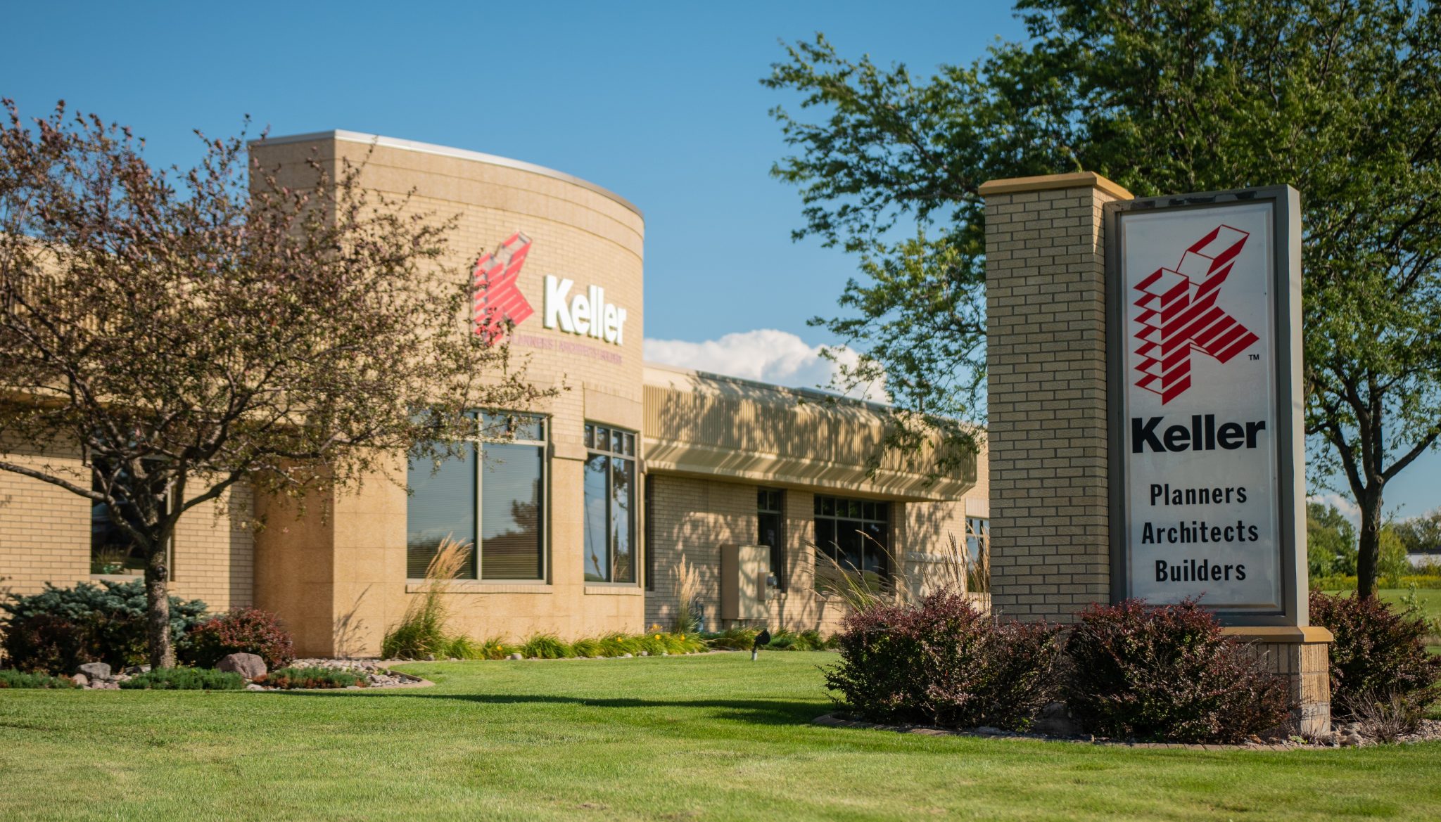 Exterior view of Keller's office building, showcasing the company logo and sign that reads "Keller Planners Architects Builders" with landscaped grounds and clear blue sky in the background.
