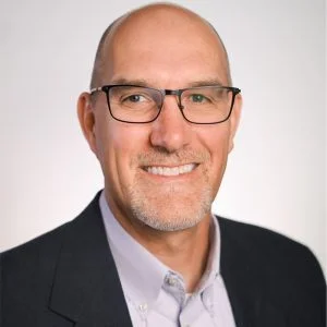 Professional headshot of a smiling middle-aged man with glasses, wearing a black blazer over a collared shirt, set against a neutral background.