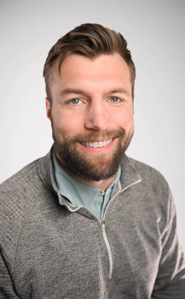 Smiling man with a beard wearing a gray zip-up sweater over a light blue shirt, against a neutral background. The image captures a friendly and approachable expression, ideal for professional profiles or personal branding.