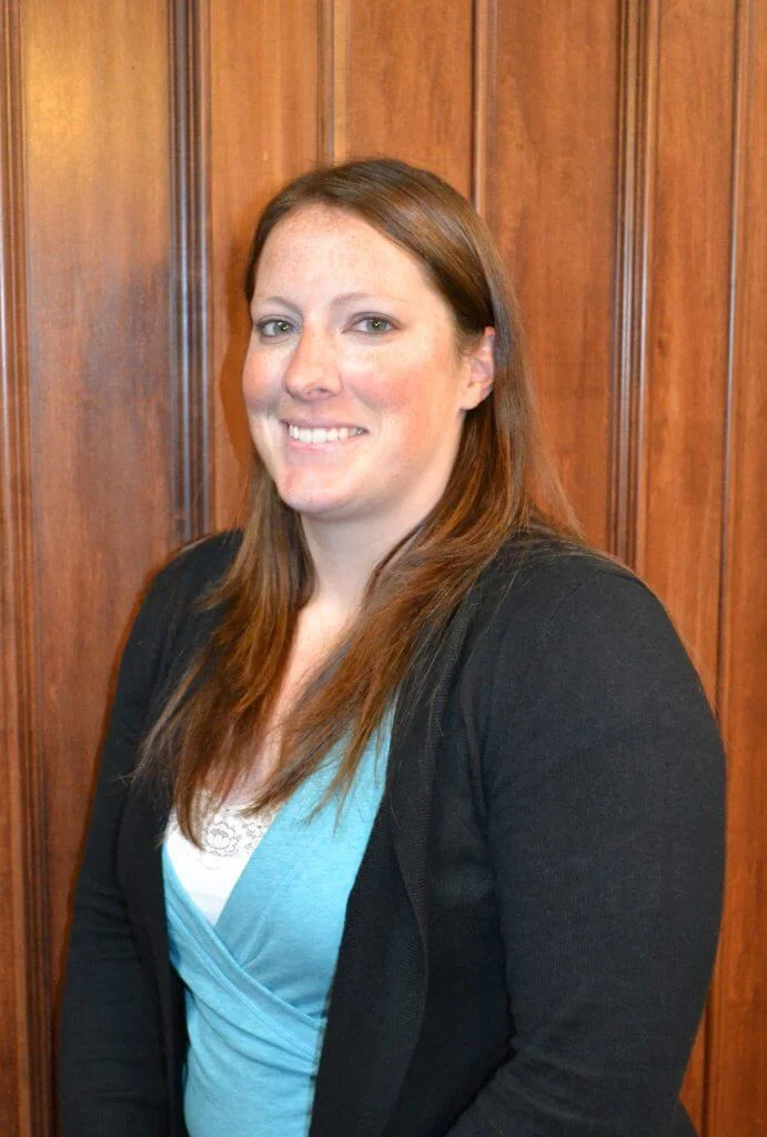 Smiling young woman with long brown hair wearing a turquoise top and black cardigan, standing against a wooden background.