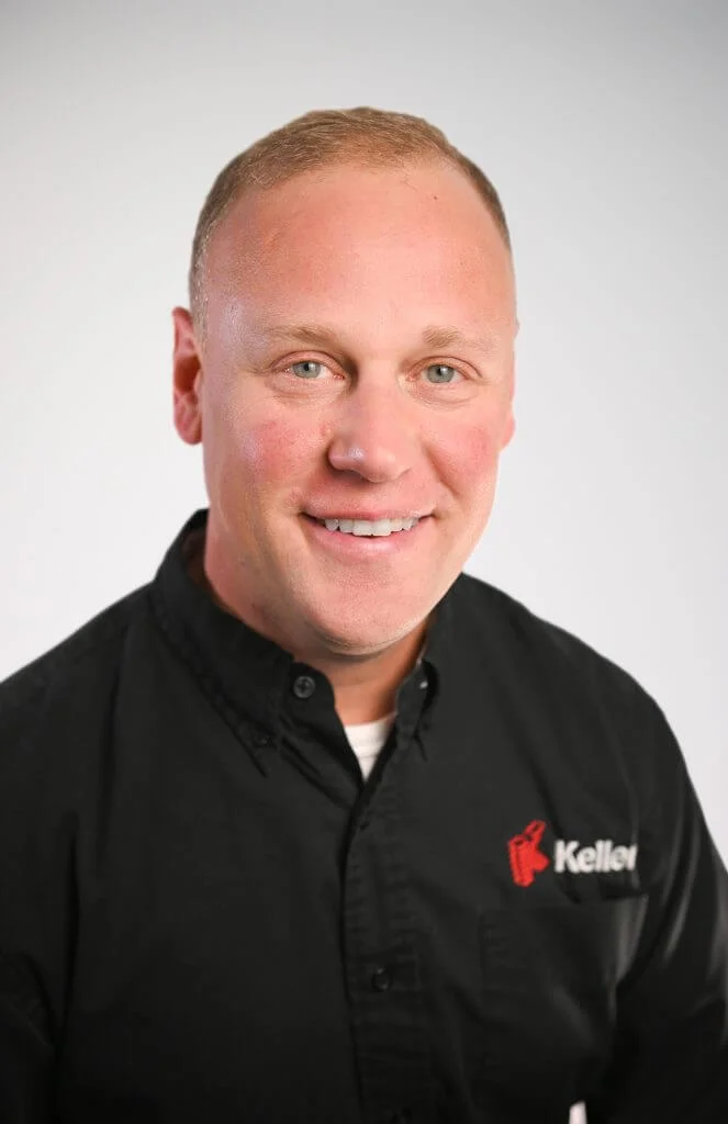 Smiling professional wearing a black shirt with a logo, posing against a neutral background, ideal for business profiles or team introductions.