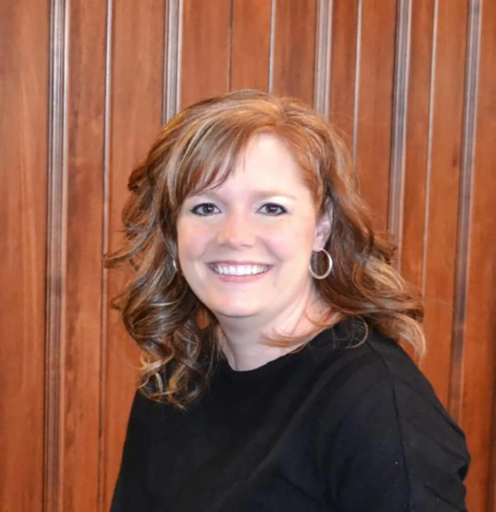 Smiling woman with curly hair wearing a black top and hoop earrings, positioned in front of a wooden panel backdrop.
