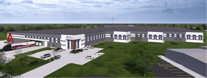 Aerial view of a modern industrial facility with a large parking area, featuring a prominent entrance with the company logo. The site includes a delivery truck and multiple vehicles, surrounded by landscaped green spaces and trees.