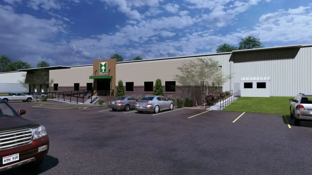 Exterior view of a modern warehouse building with a landscaped parking lot, featuring several parked cars and a clear blue sky in the background. Signs and ramps indicate accessibility, showcasing a welcoming entrance.